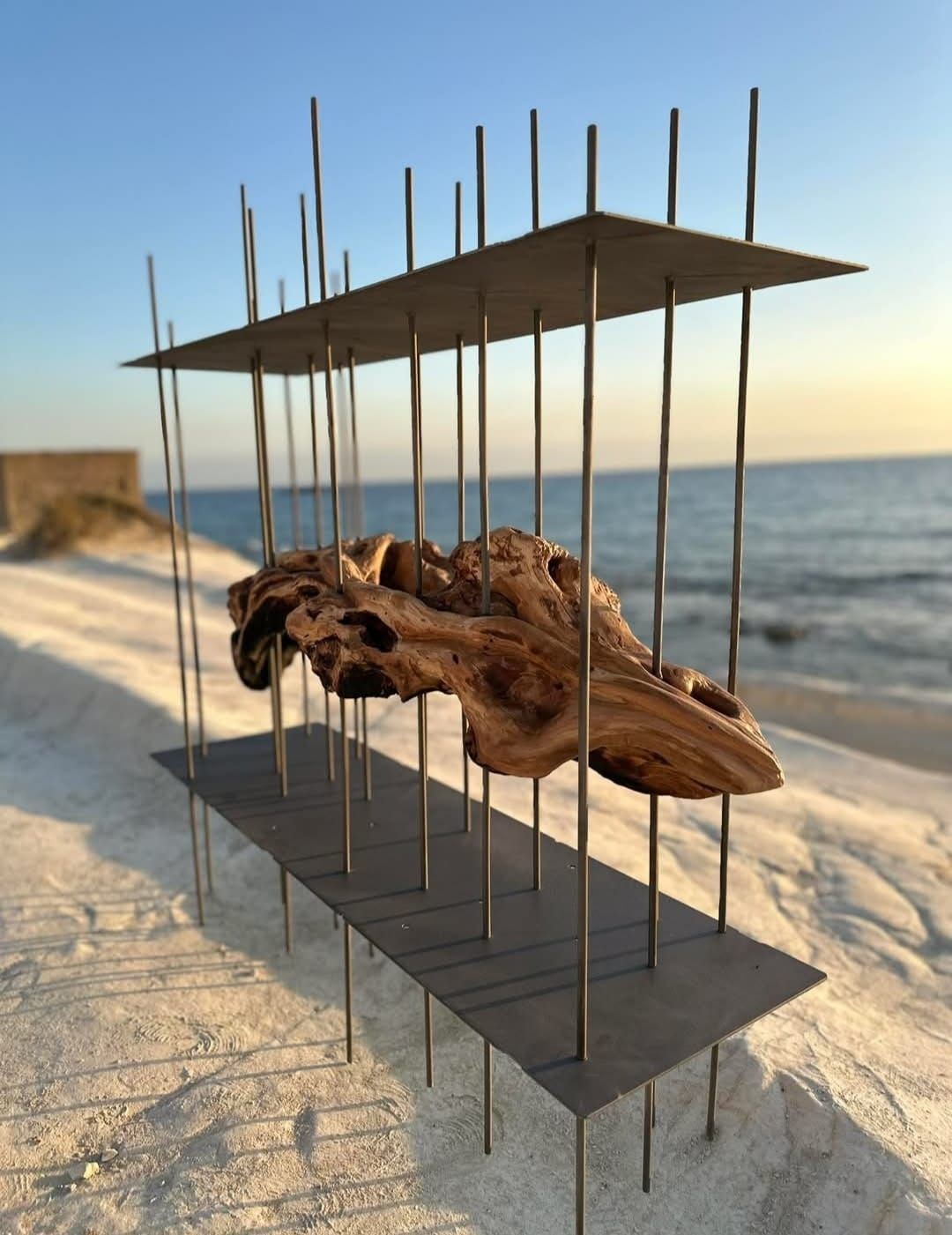 Legno galleggiante scultoreo racchiuso da barre e piastre metalliche su una spiaggia sabbiosa.