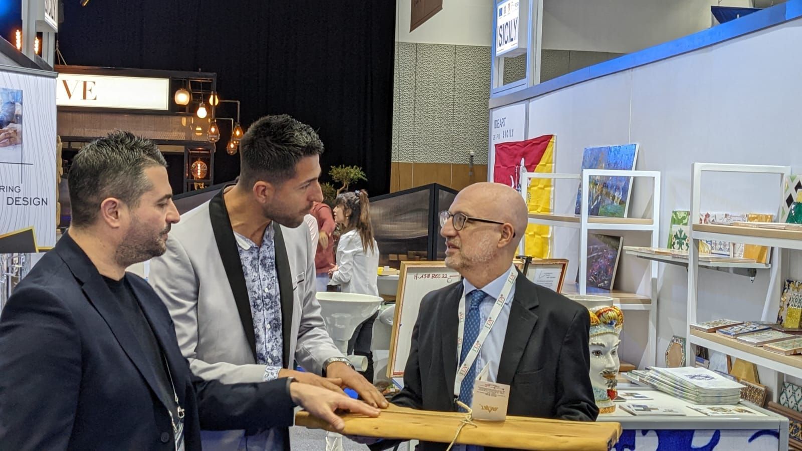 Tre uomini in uno stand fieristico discutono di prodotti. Uno indica un espositore, gli altri osservano.