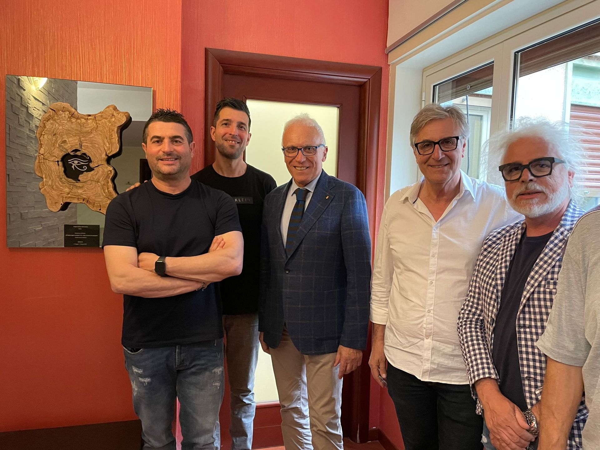 Gruppo di uomini in piedi all'interno di una stanza con un quadro incorniciato. Tre sono in giacca e cravatta, gli altri in abiti casual.