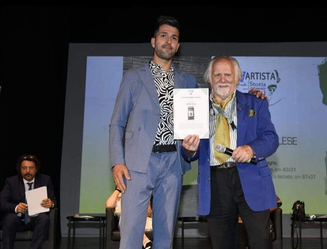 Due uomini sul palco con in mano un certificato. Uno indossa un abito blu, l'altro un blazer. Un uomo in abito è seduto.