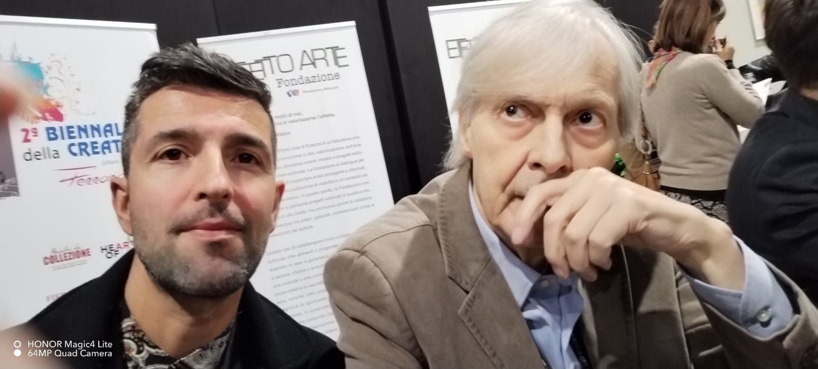 Due uomini, uno con i capelli grigi e una giacca, l'altro con la barba scura, posano vicino a un poster bianco.