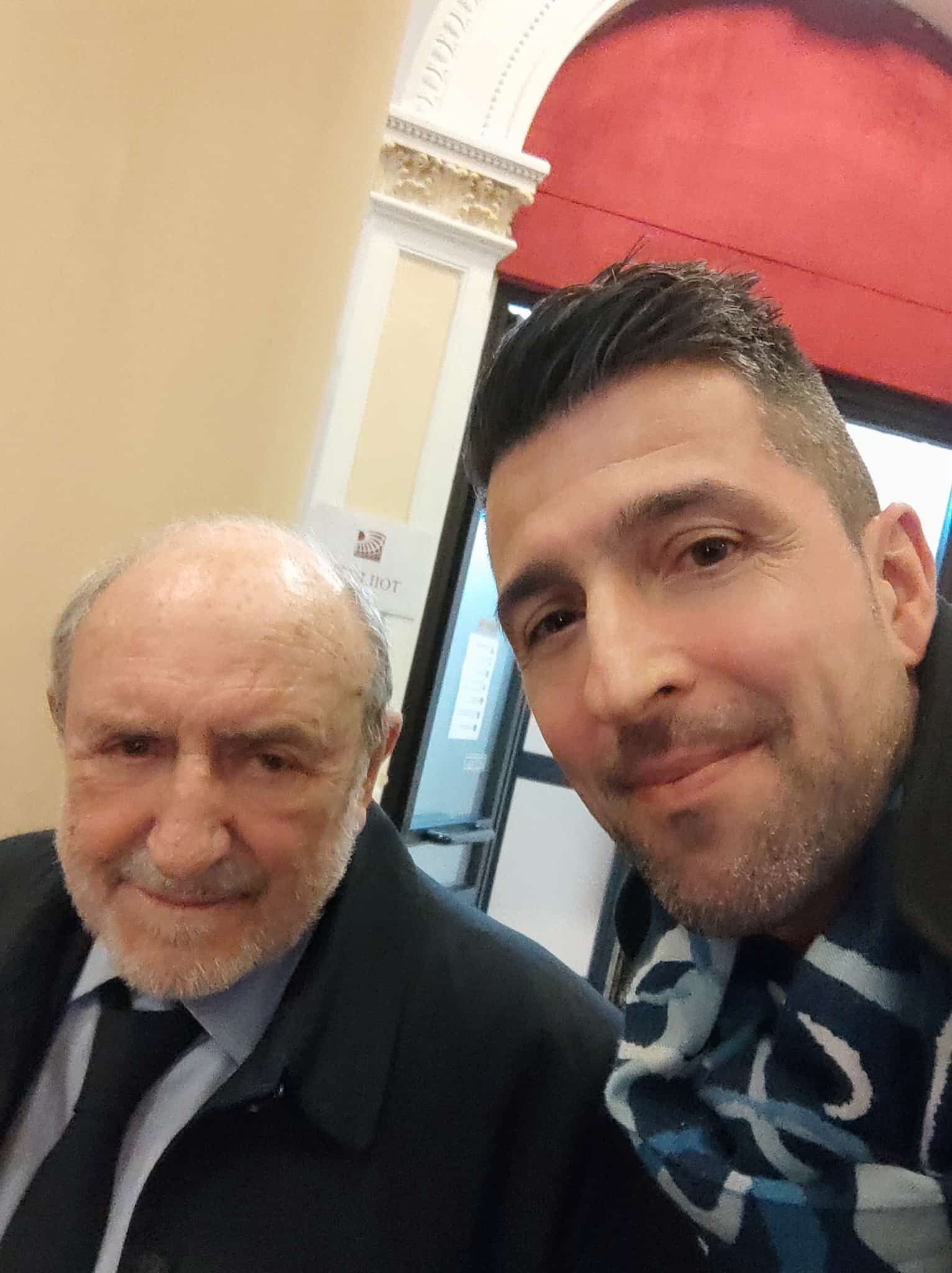 Un uomo si scatta un selfie con un uomo anziano; entrambi sorridono all'interno di un edificio.