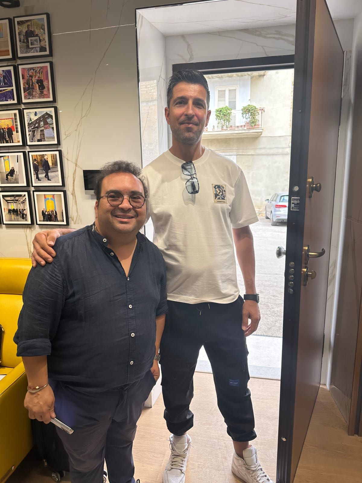 Due uomini sono in piedi dietro una porta. Uno ha un braccio intorno alle spalle dell'altro. Stanno sorridendo.