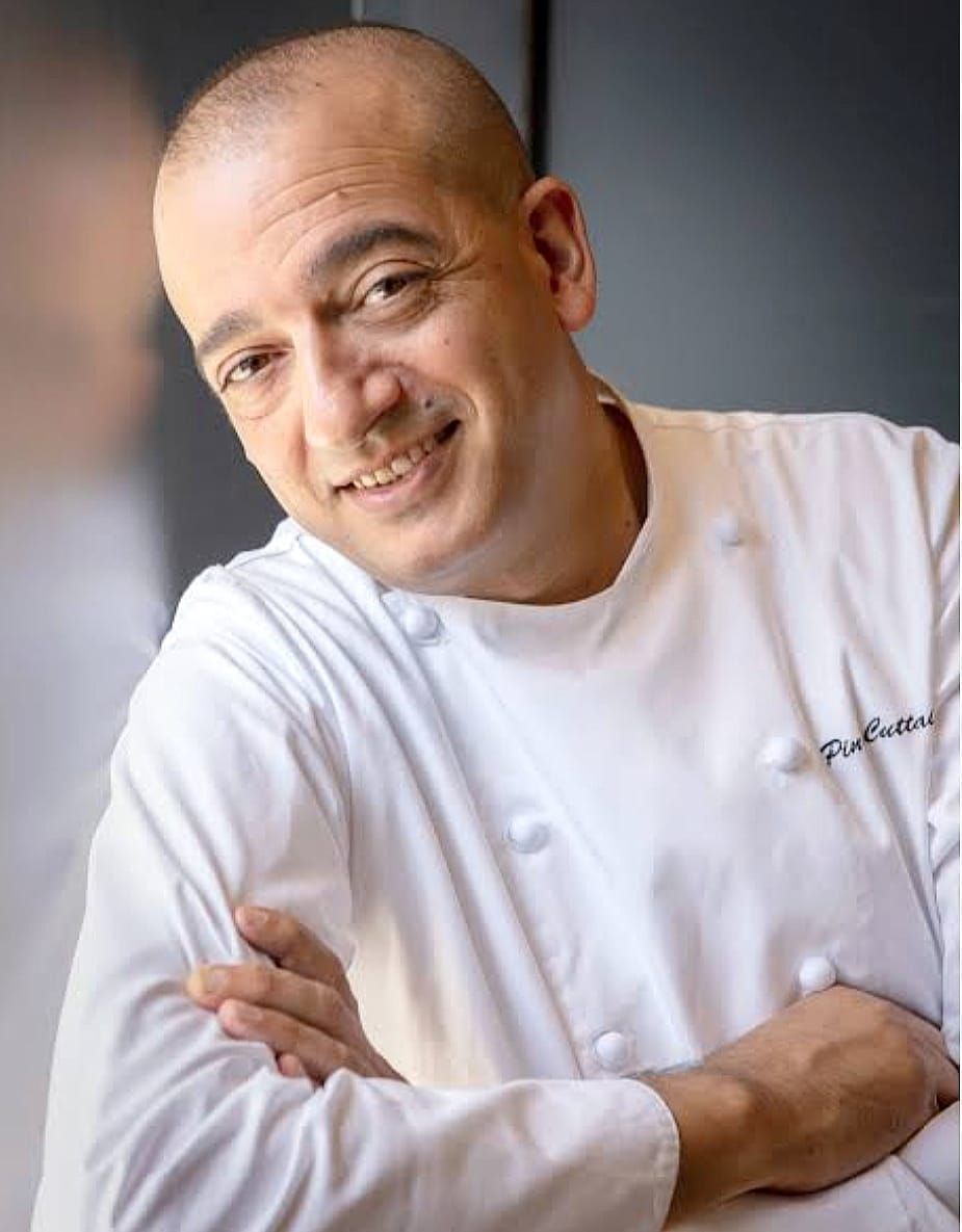 Cuoco in camice bianco, sorridente, braccia incrociate, appoggiato a una superficie riflettente.