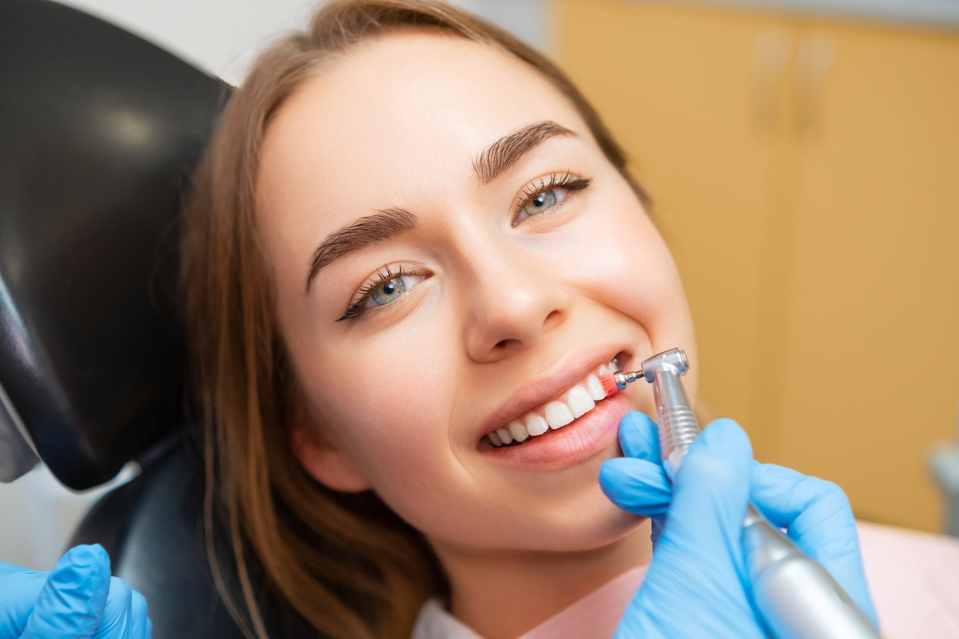 Blue-gloved hands are performing teeth cleaning on a young woman’s bright smile.