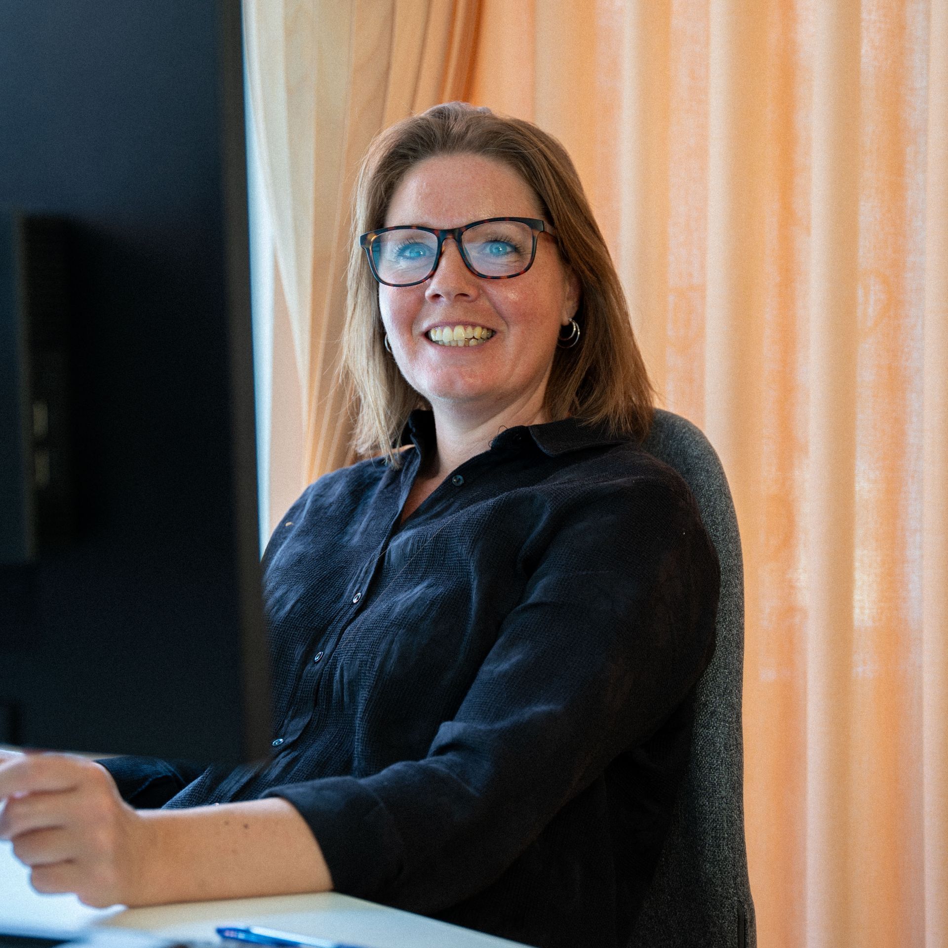 Eine Frau mit Brille sitzt lächelt an einem Schreibtisch vor einem Computermonitor.