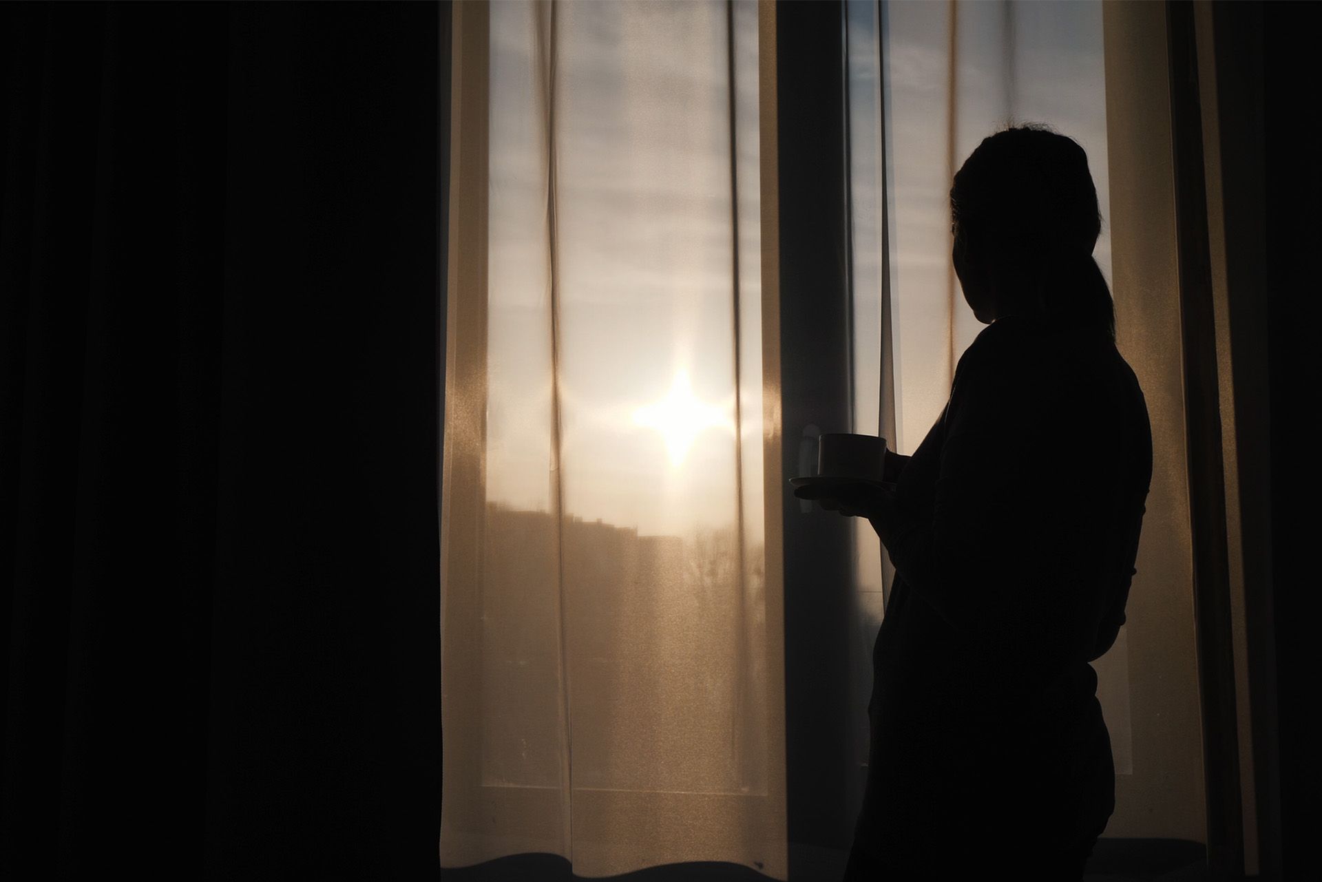 A woman is standing in front of a window looking out at the sunset.