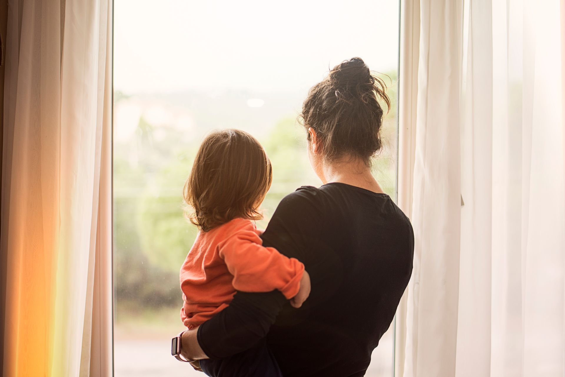 A woman is holding a child and looking out of a window.