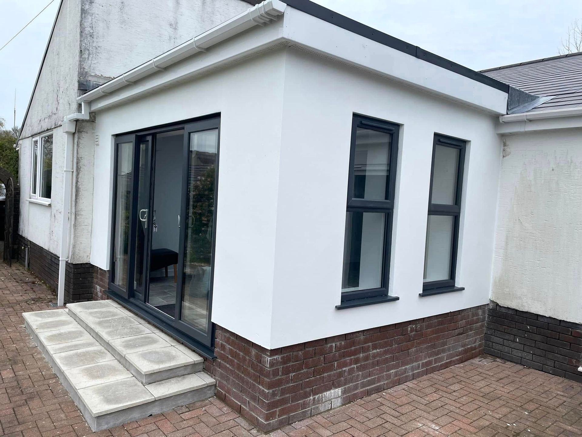 A newly built extension on a house, featuring gray stucco walls, brick base, dark-framed windows, and sliding glass doors.
