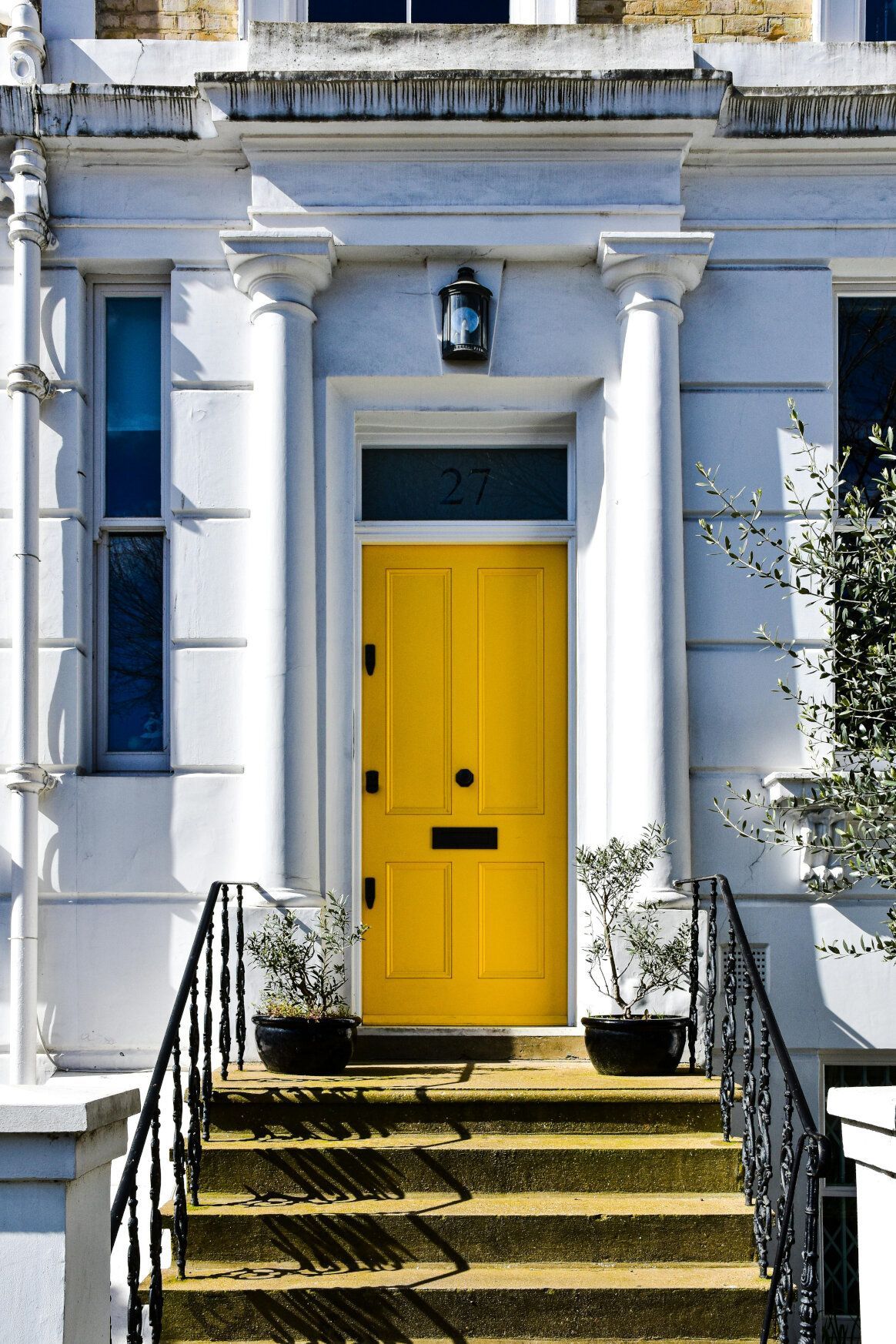 Yellow door on a white building, flanked by columns and steps with black railings.