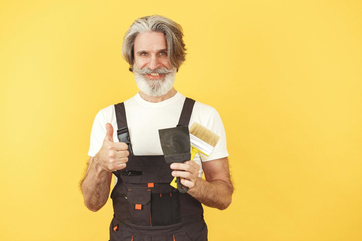Man with gray hair and beard in overalls gives a thumbs up, holding a scraper and brush, yellow background.