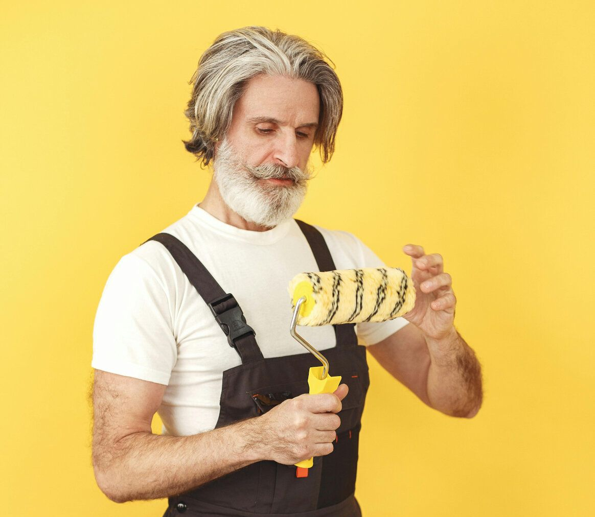 Senior painter in overalls examining a paint roller against a yellow background.