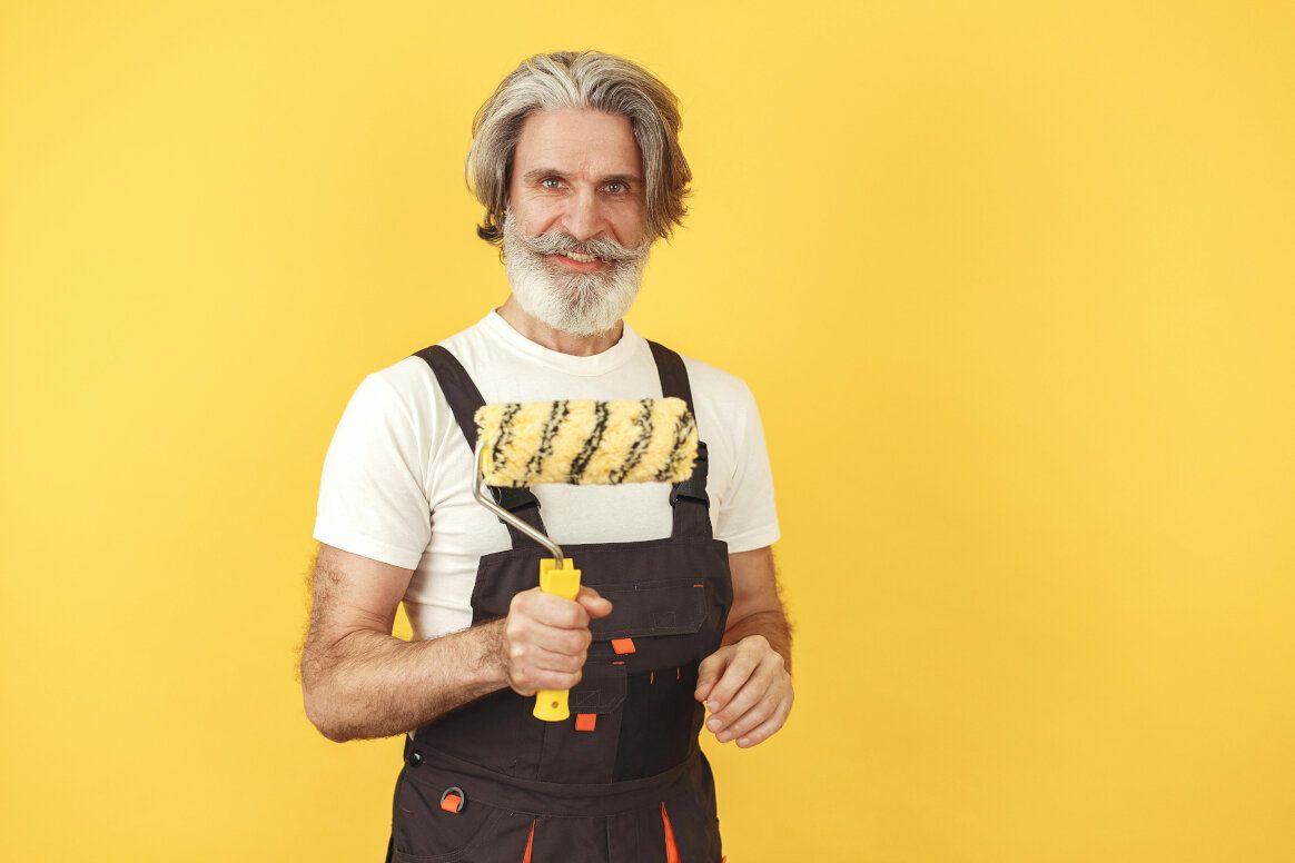 Smiling older man holding a paint roller on a yellow background. He wears overalls and a white shirt.