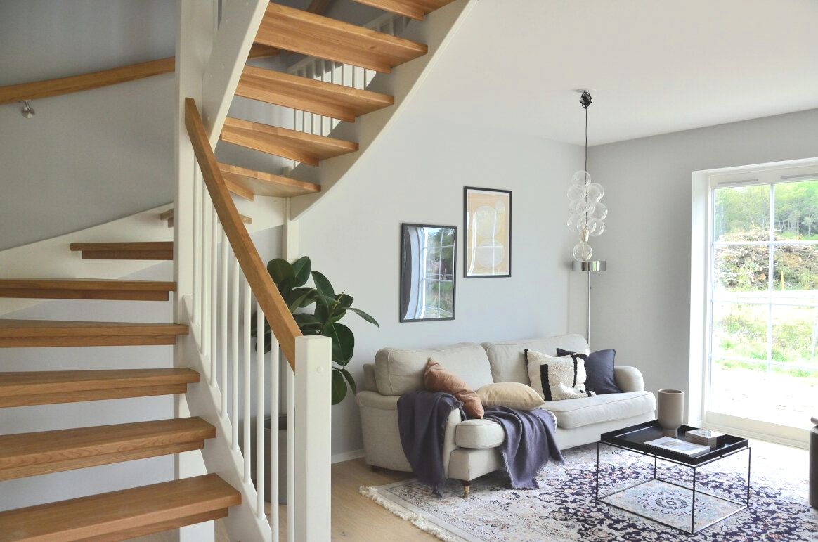 Living room with wooden stairs, sofa, and a rug. Soft colors and natural light create a cozy atmosphere.