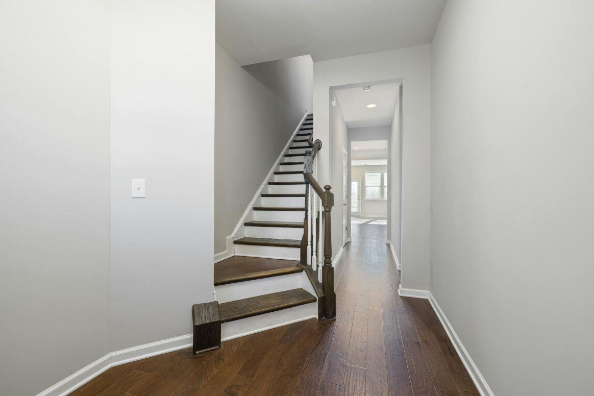 Hallway with dark wood floor, stairs with brown carpet, and a view into a room with a window.