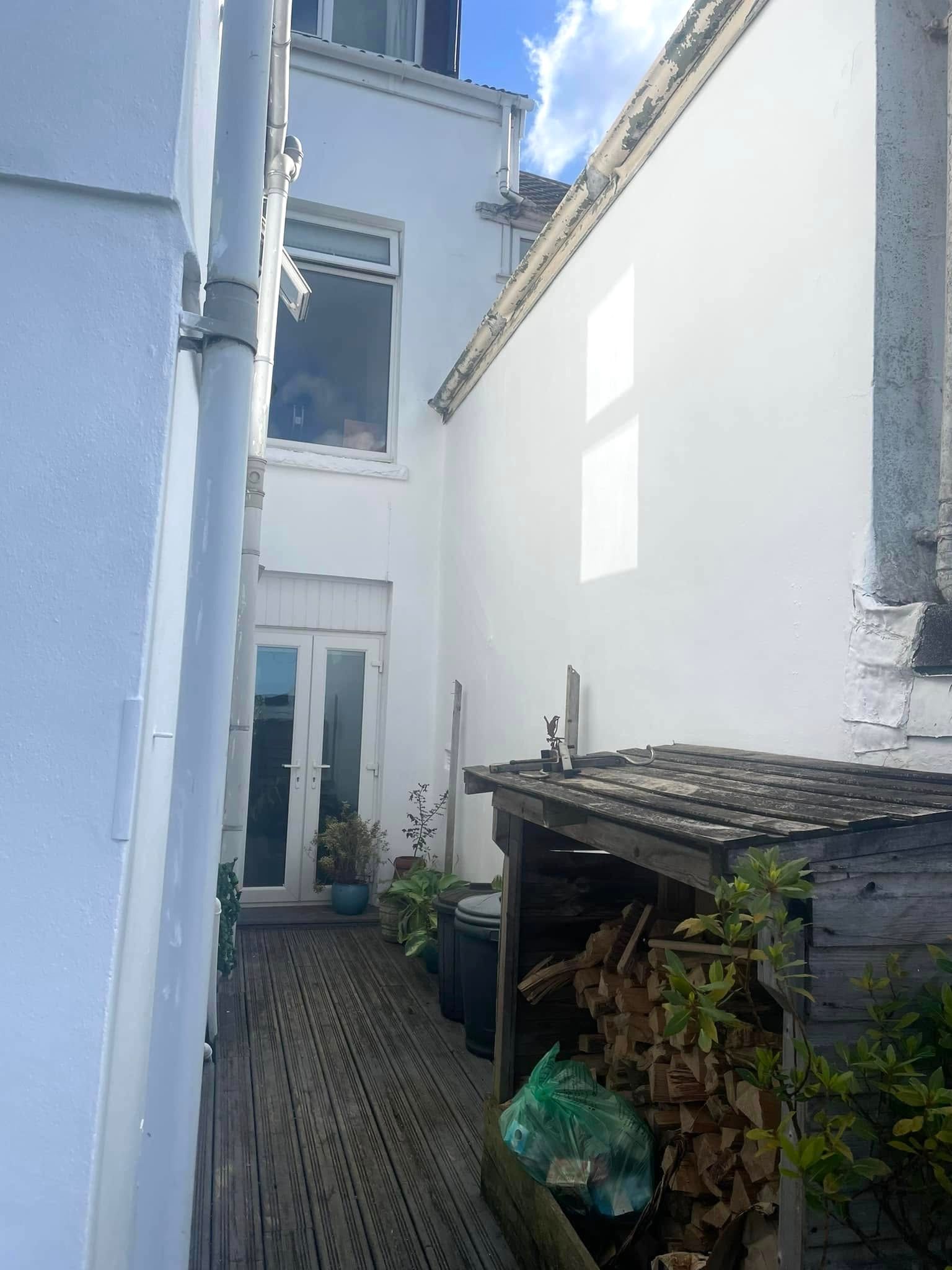 Narrow outdoor space between white buildings; wooden walkway, firewood shed.