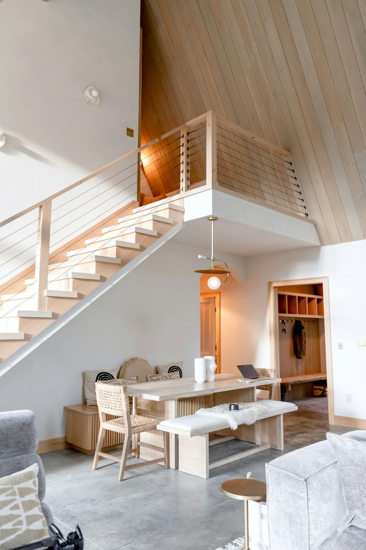 Bright, modern interior with stairs to a loft. Wooden dining table, concrete floor, and light wood walls.