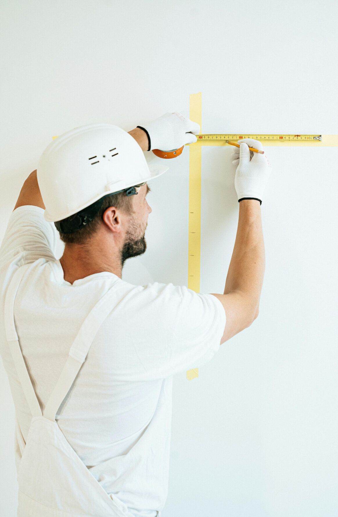 Man in white overalls and hard hat using a ruler to measure yellow tape on a white wall.