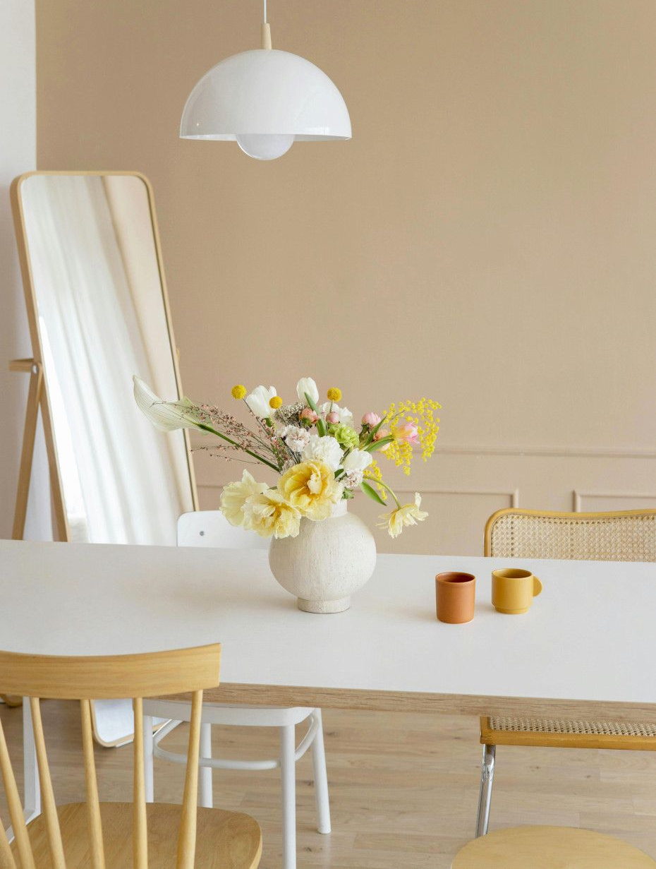 Dining room with white table, vase of flowers, and yellow chairs. Light-colored walls, mirror, and pendant light.