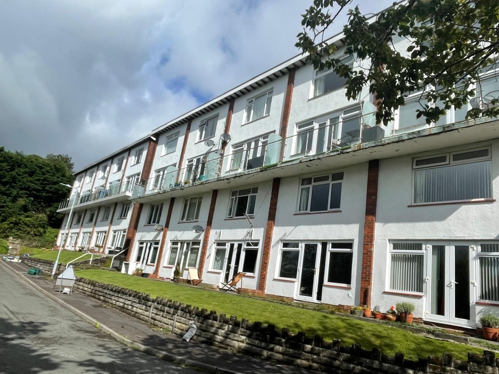 White multi-story apartment building with balconies, set on a sloping green hill, under a cloudy sky.