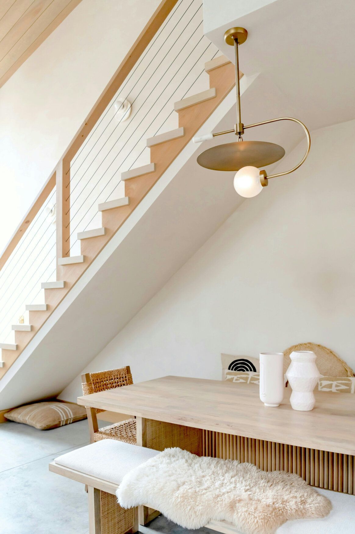 Wooden dining table and bench beneath a staircase, with a modern light fixture and minimalist decor.