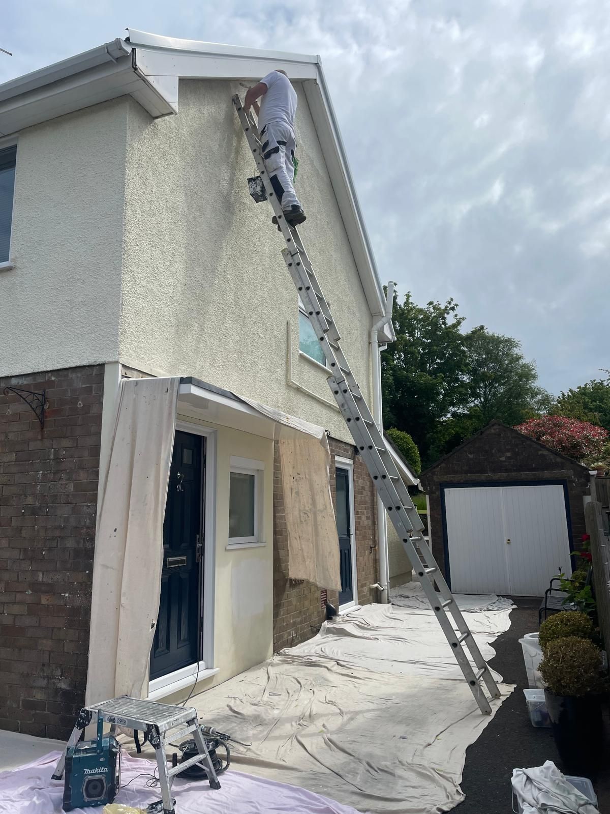 Person painting house exterior from a tall ladder. Cloudy sky.