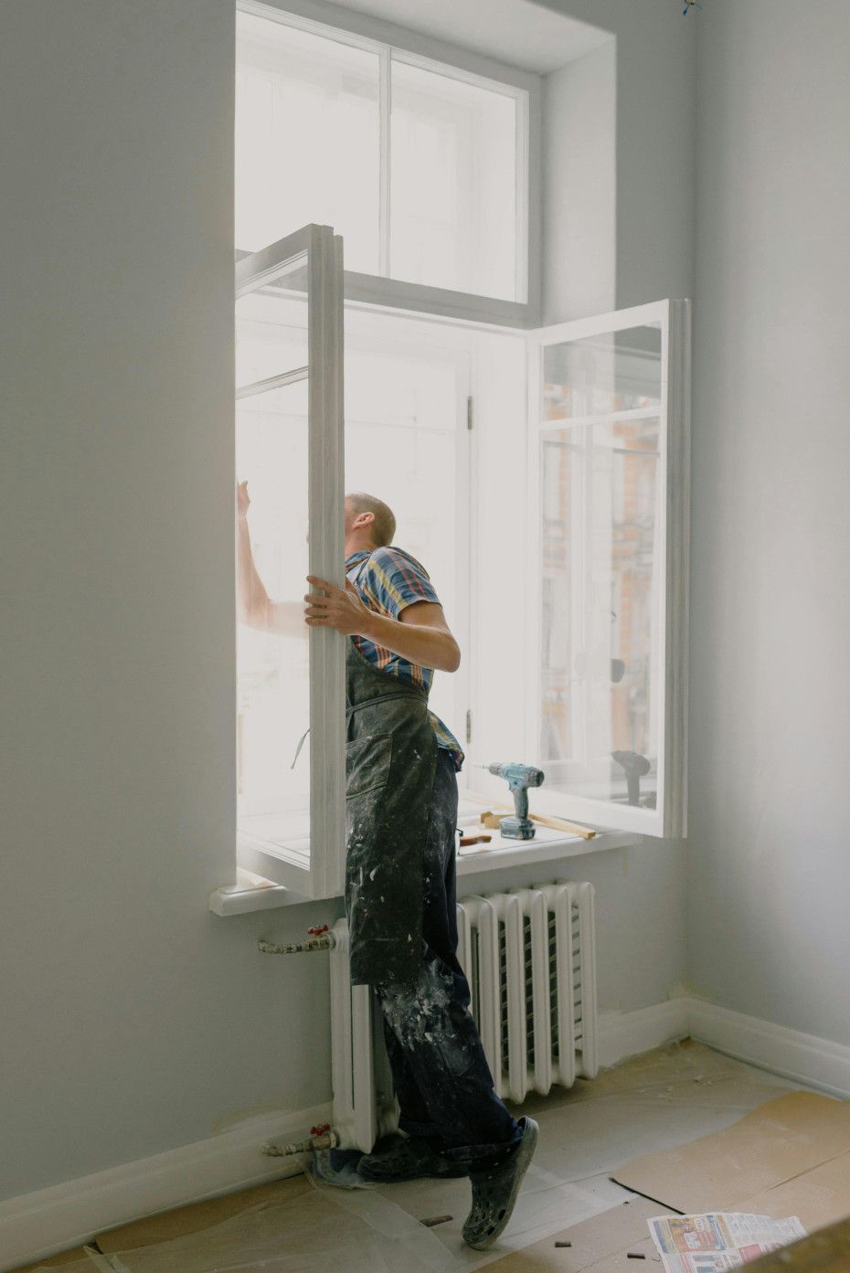 Man installing a white window frame indoors. He is wearing overalls, standing near the window, with tools on the windowsill.