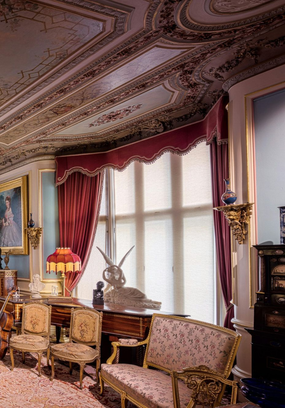 Ornate room with red curtains framing a window with a rabbit statue. Gold furniture and detailed ceiling.