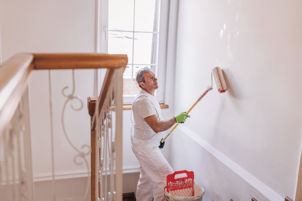 Decorator painting a white wall with a roller in Swansea home. Bucket of paint is on the stairs, next to the wall. By Daffodil Decor.