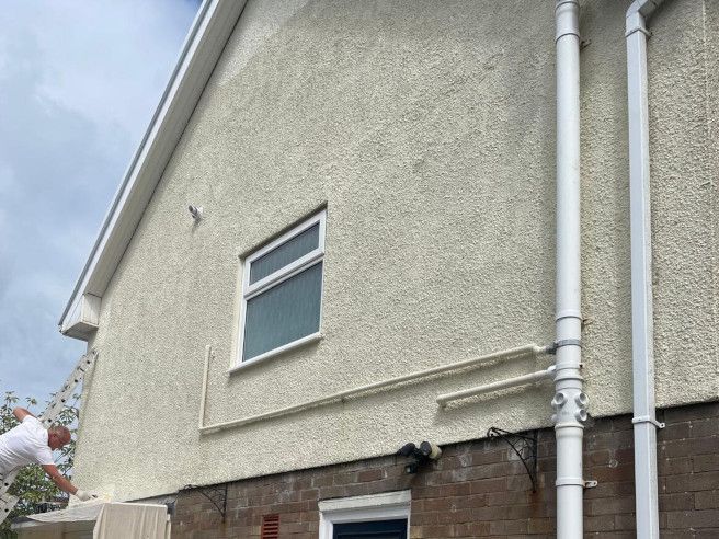 Expert exterior painter on ladder painting external render pebbledash on a terraced semi-detached Swansea house in cream render paint on a cloudy day by Daffodil Decor - close up shot.