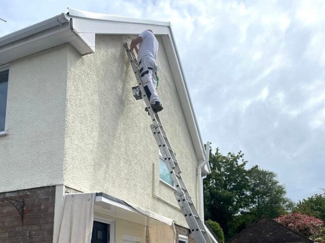 Expert exterior painter on ladder painting external render pebbledash on a terraced semi-detached Swansea house in cream render paint on a cloudy day by Daffodil Decor.