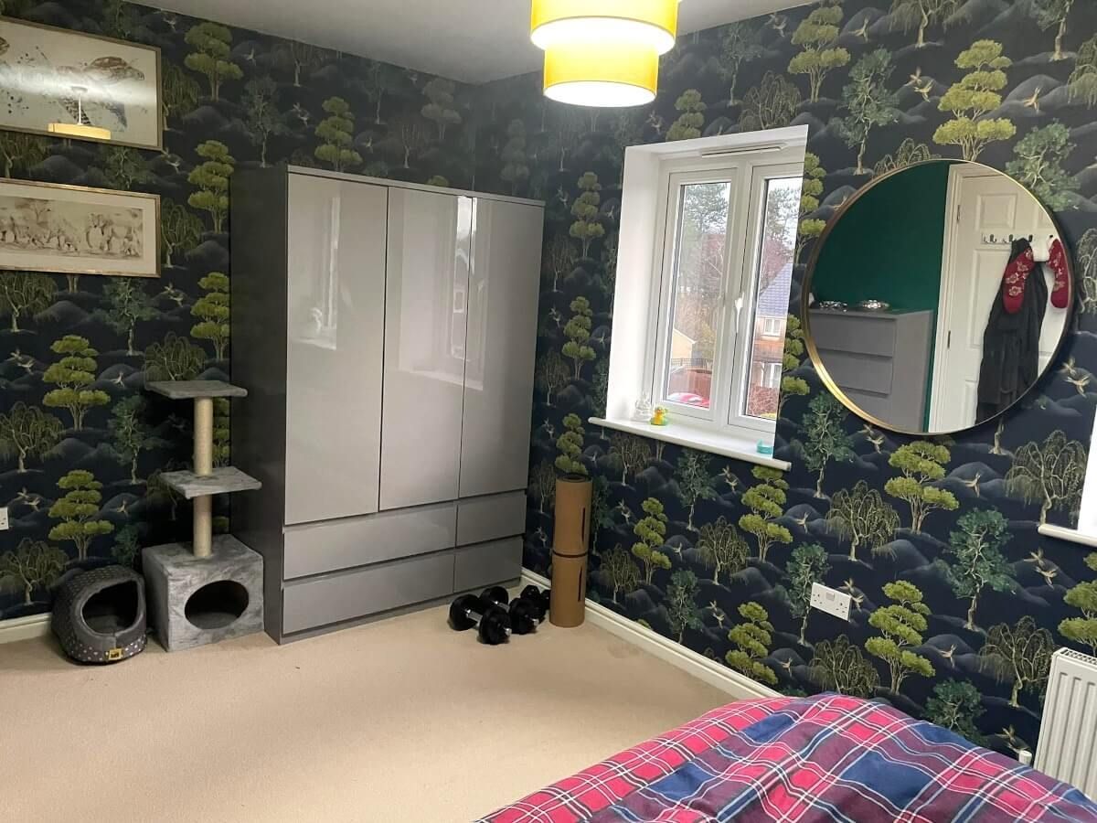 A Swansea bedroom with green and blue patterned wallpaper installed by Daffodil Decor, a gray wardrobe, cat tree, round mirror, and a window.
