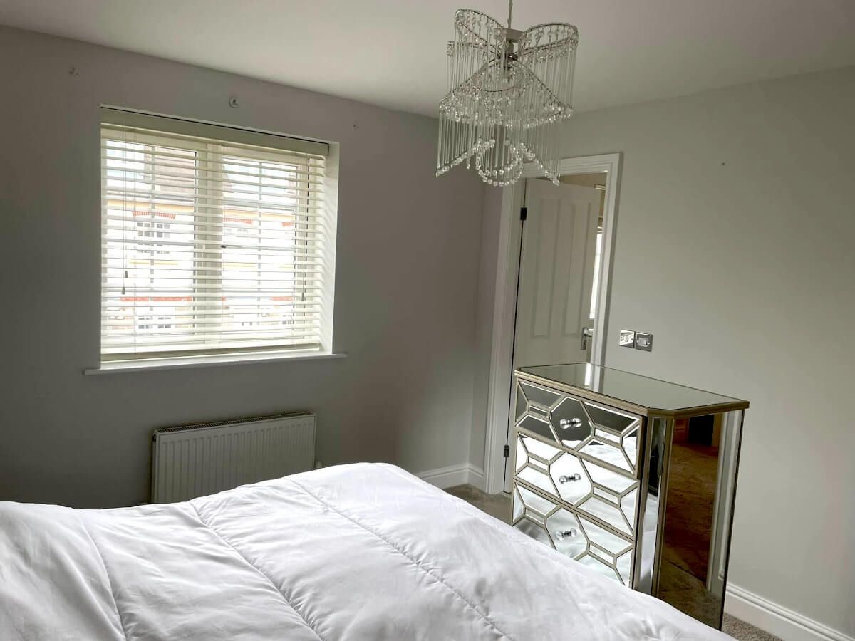 Bedroom with a bed, mirrored dresser, window with blinds, and chandelier. Gray walls painted by Daffodil Decor in Swansea.