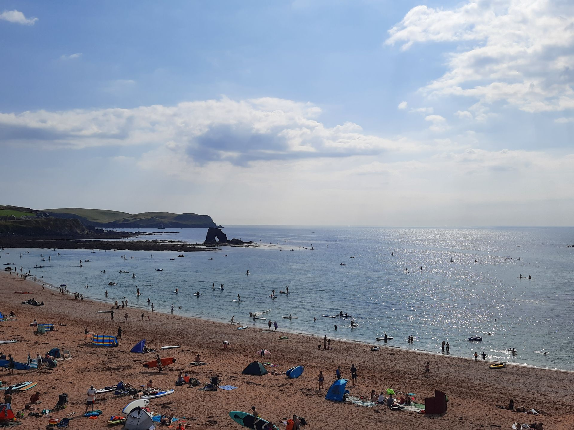 Sandy beach with blue sea, people swimming and paddleboarding, brown cliffs, and green hills under a blue sky.