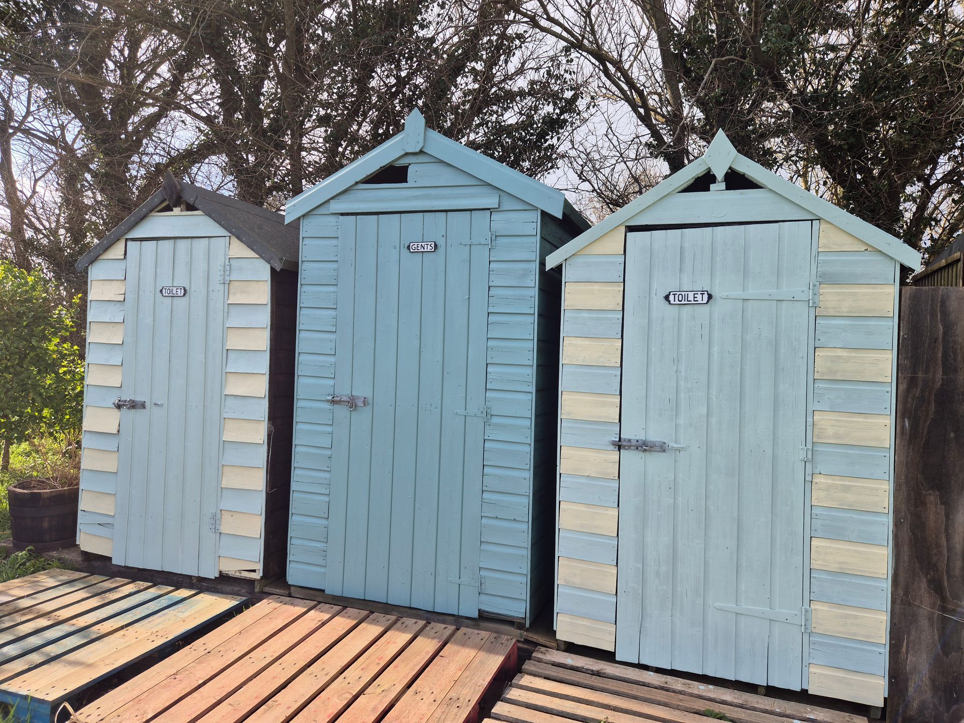 Three wooden beach huts with blue and yellow stripes. Blue sky and green trees behind. 