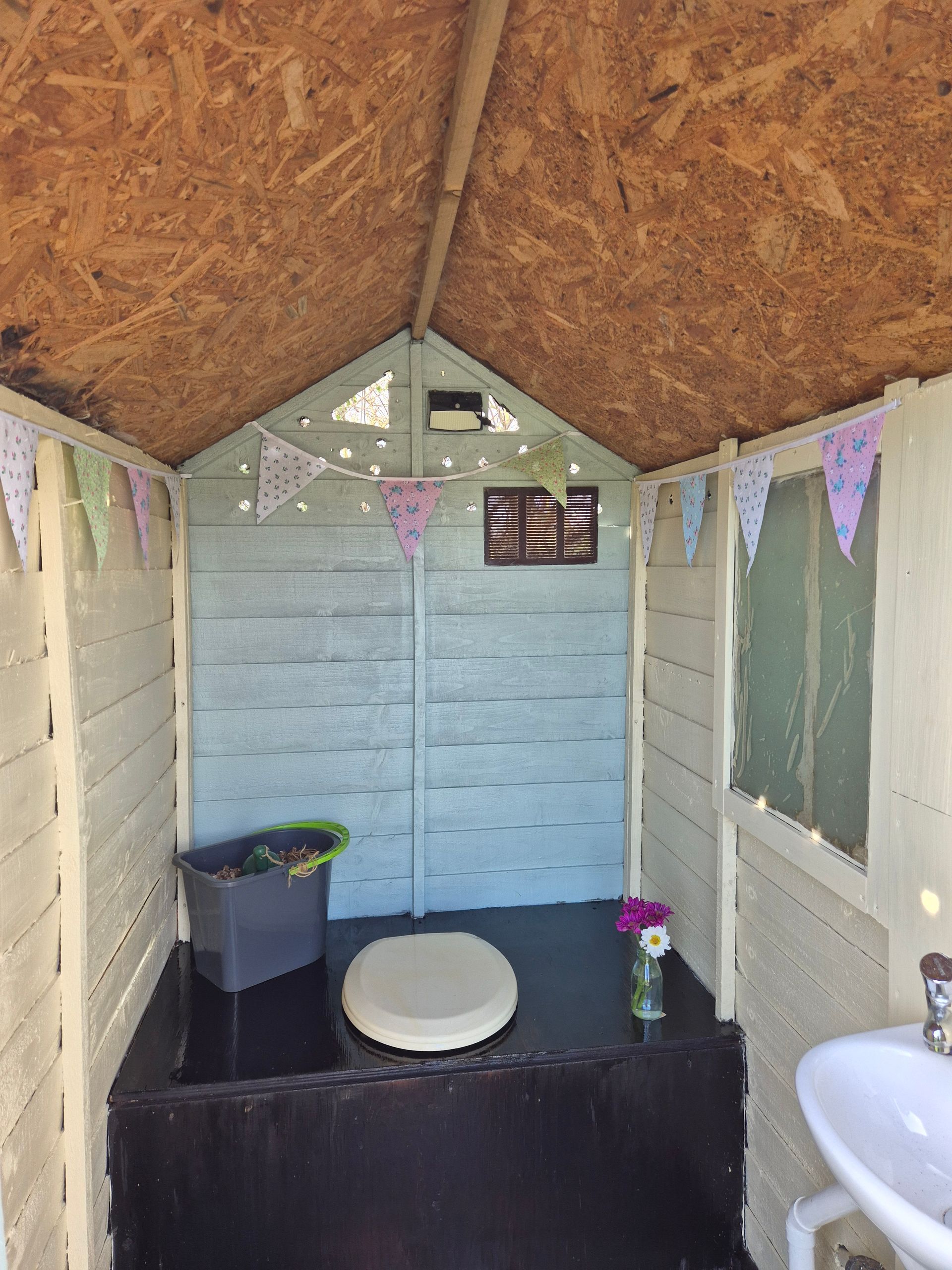 Blue and cream wooden shed, with white loo and sink and floral vintage bunting.