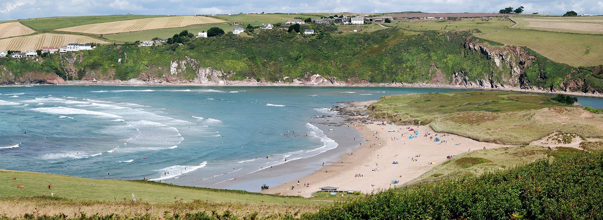 Coastal bay with a sandy beach, green hills, and blue water under a cloudy sky.