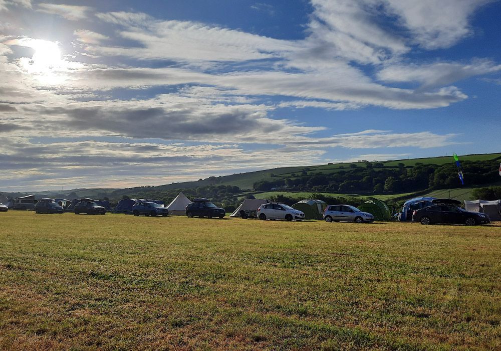 Camping field with tents and cars under a partly cloudy sky and bright sun, with green hills in the background.
