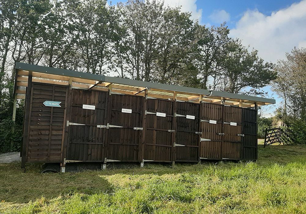Wooden outdoor restroom stalls with a roof, set in a grassy area with trees and a blue sky.