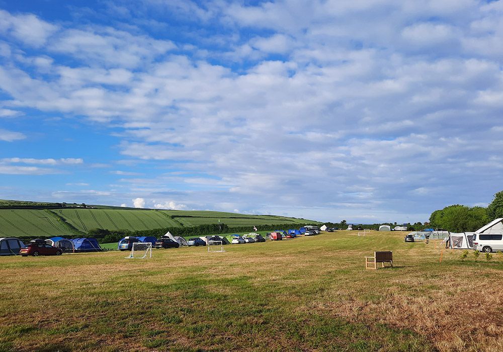 Campsite with tents in a grassy field under a partly cloudy blue sky. Rolling green hills in the background.