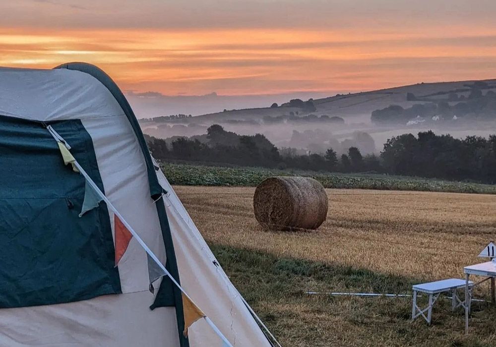 Camping tent in a field at sunrise with a hay bale and misty hills in the background.