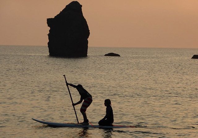 Silhouette of people paddleboarding on calm water at sunset, with a large rock formation in the background.