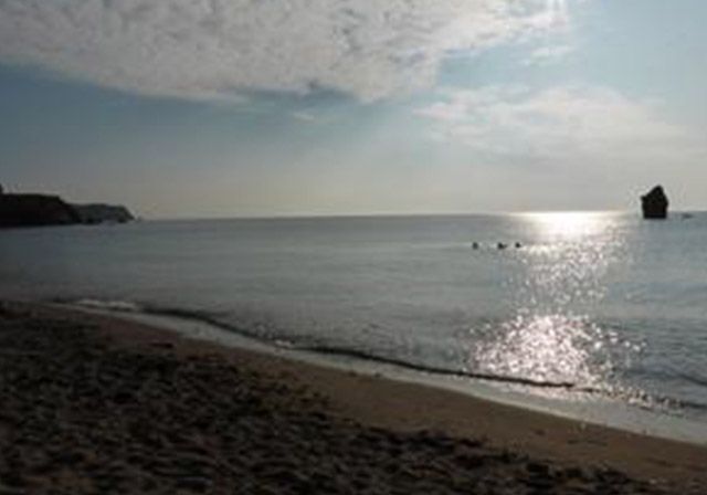 Beach scene at daytime with sun reflecting on water, small waves, sandy shore and rock formations.