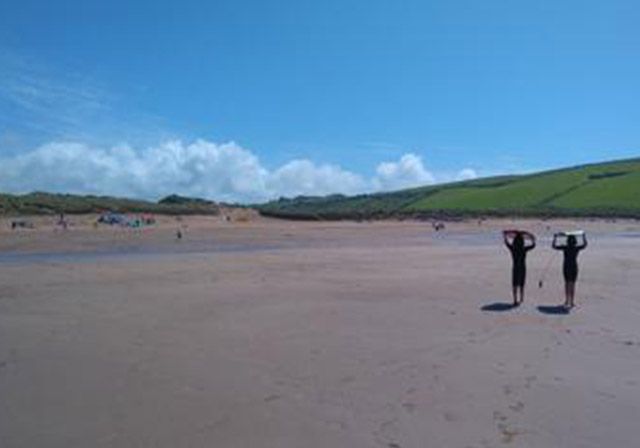Two people on a sandy beach holding surfboards on a sunny day. Green hills and blue sky in the background.