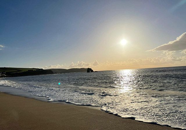 Beach scene with sun reflecting on the ocean's surface. Waves meet the shore under a clear sky.