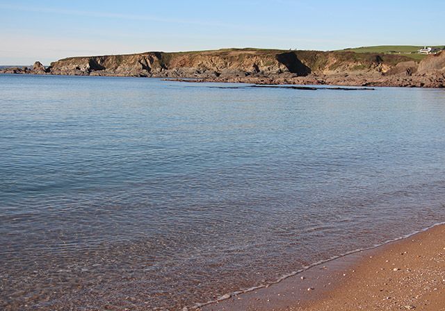 Calm ocean water meets a sandy beach, with a rocky cliff in the background under a blue sky.