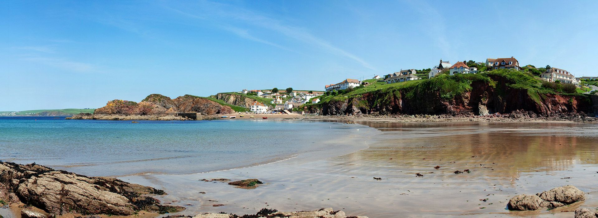 Coastal landscape with buildings on a green cliff, calm blue sea, and a sandy beach under a clear sky.