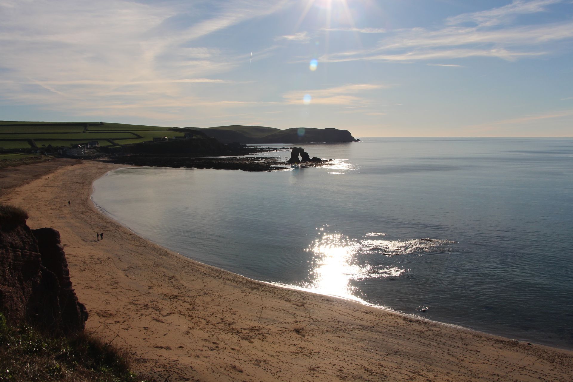 Sandy beach with sea, brown cliffs, green hills under a blue sky and sunshine.