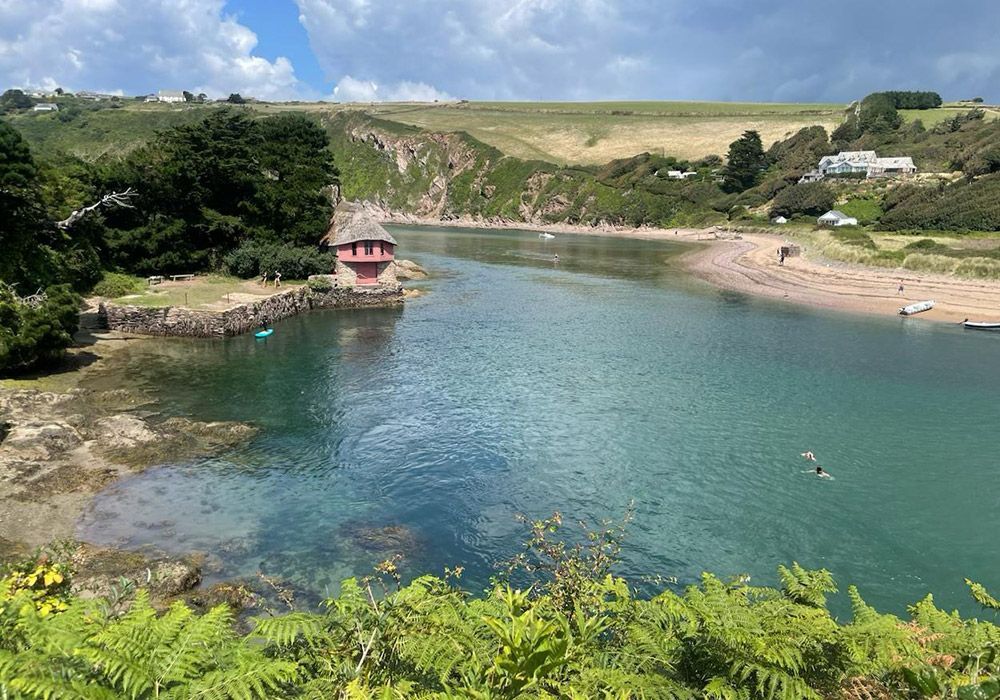 A scenic cove with calm, turquoise water. A small, red building sits on a rocky edge; green hills in the background.