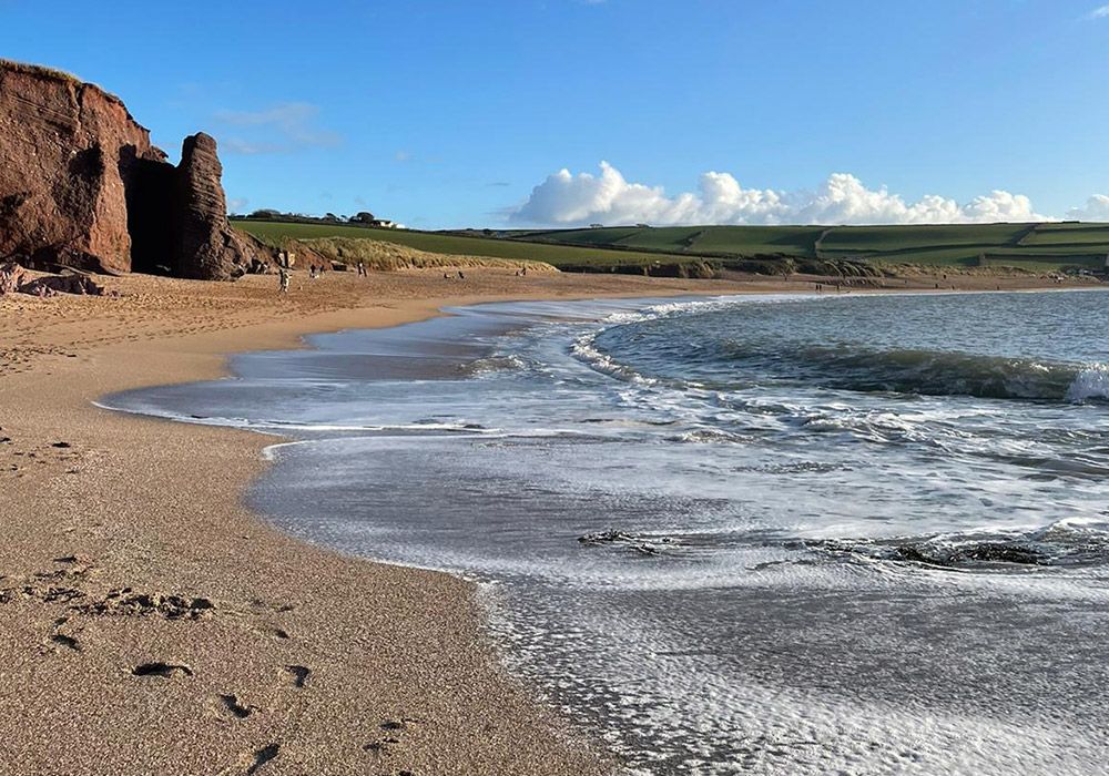 Sandy beach with waves, red cliffs, and green hills under a blue sky.